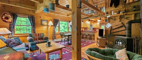 Een gezellig blokhutinterieur met een eethoek, een bank, een open haard en een keuken die door een open deur zichtbaar is.