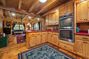 Interior de una cabaña con pisos de madera, una cocina con armarios de madera, una zona de comedor con una mesa roja y una chimenea.