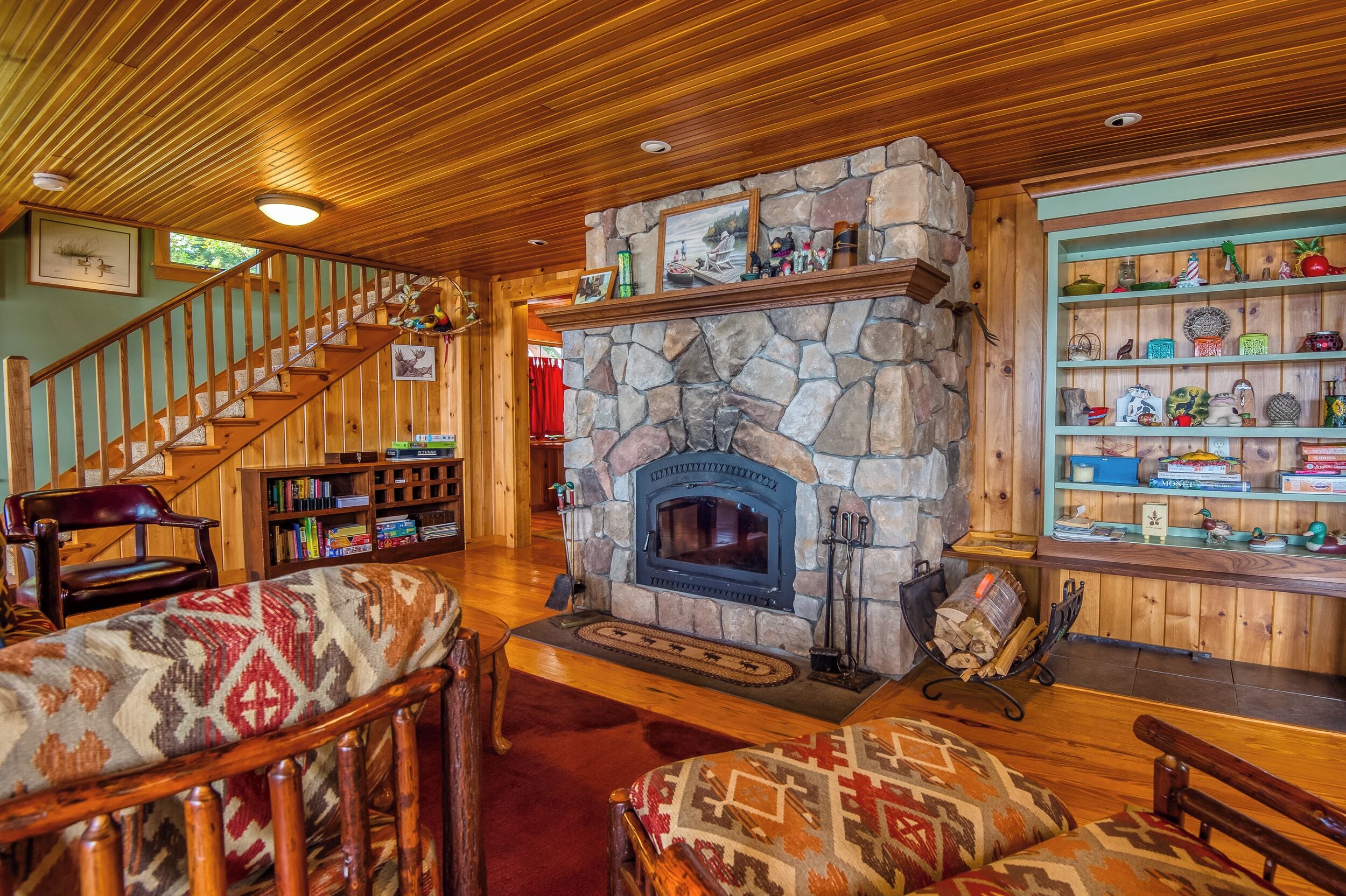 A cozy living room with a stone fireplace, wooden staircase, and a bookshelf filled with books and decorative items.