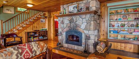 A cozy living room with a stone fireplace, wooden staircase, and a bookshelf filled with books and decorative items.