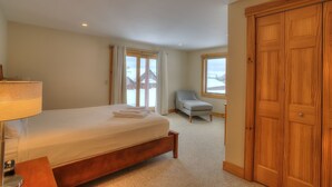 A bedroom with a bed, a chair, a wooden wardrobe, and a window with a view of a building outside.