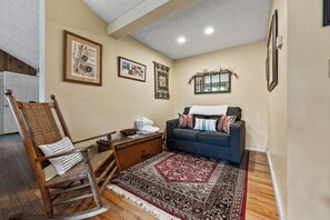 A cozy living room with a rocking chair, a sofa, a patterned rug, and framed pictures on the wall.