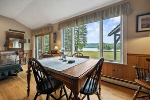 A dining area with a wooden table and chairs, large windows with a view of a lake and trees, and a cozy interior with a couch and a lamp.