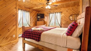 A wooden cabin bedroom with a bed, a ceiling fan, a television, and two windows with curtains. - Hart | Beautiful family getaway located just three miles from downtown Rangeley (Rangeley)