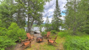 A rustic outdoor seating area with Adirondack chairs and a fire pit, surrounded by dense forest and a lake view. - Gerstley - Quiet, secluded adorable camp on Loon Lake (Rangeley)