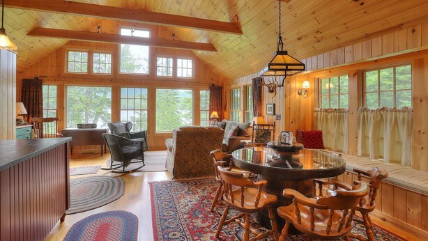 A spacious wooden cabin with a high ceiling, large windows, a round dining table, and a cozy seating area. - Gerstley - Quiet, secluded adorable camp on Loon Lake (Rangeley)