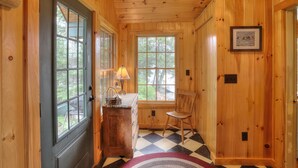 A wooden cabin interior with a checkered floor, a wooden door, a window with a view of trees, a lamp, and a chair. - Gerstley - Quiet, secluded adorable camp on Loon Lake (Rangeley)