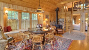 A cozy cabin interior with a wooden interior, a round dining table with chairs, a fireplace, and a large window with curtains. - Gerstley - Quiet, secluded adorable camp on Loon Lake (Rangeley)