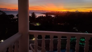 Un balcón con vista a la puesta de sol sobre el océano, palmeras y edificios a lo lejos.