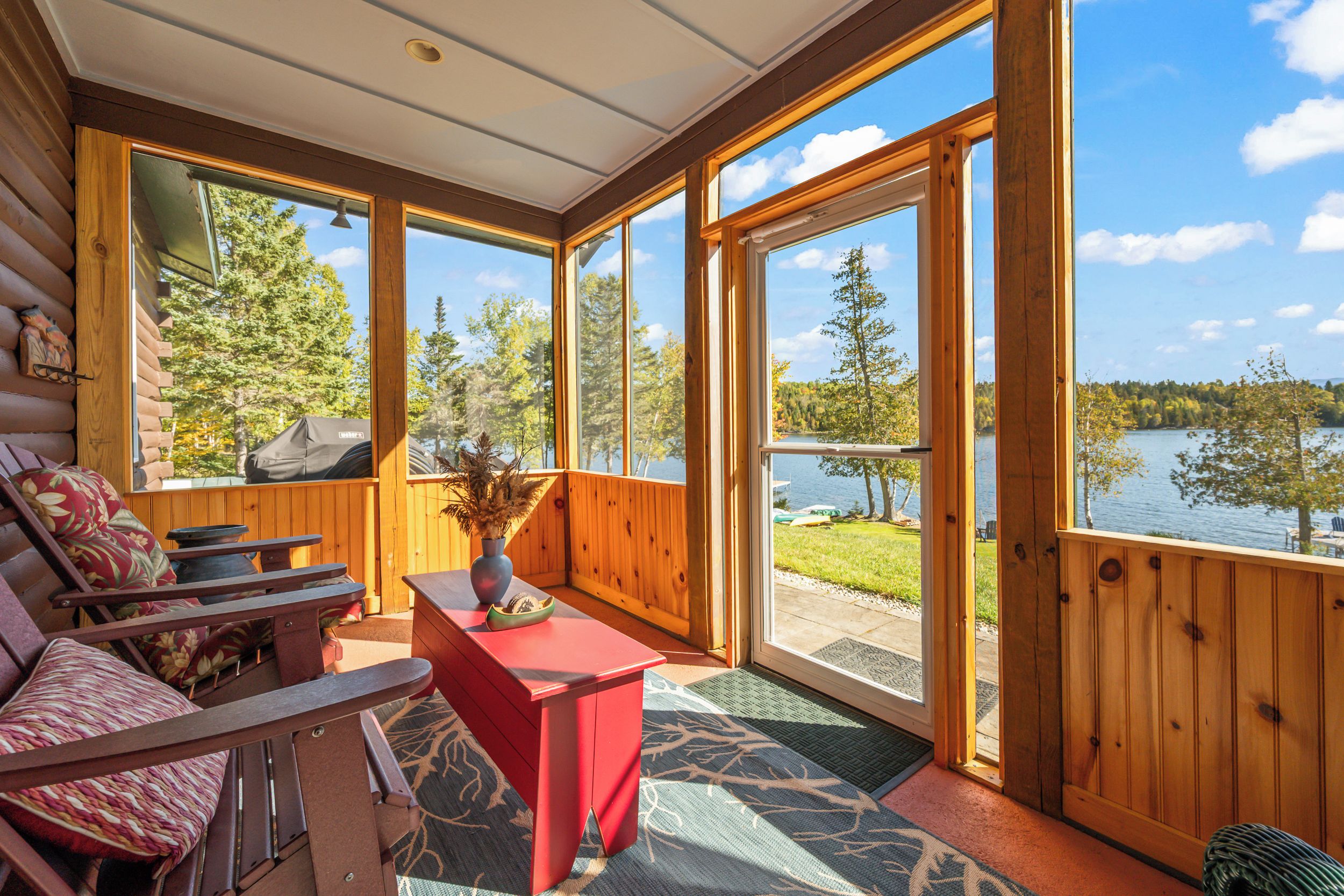 A cabin with a large sliding glass door leading to a lakeside view. Inside, there is a wooden bench and a small table with a vase.