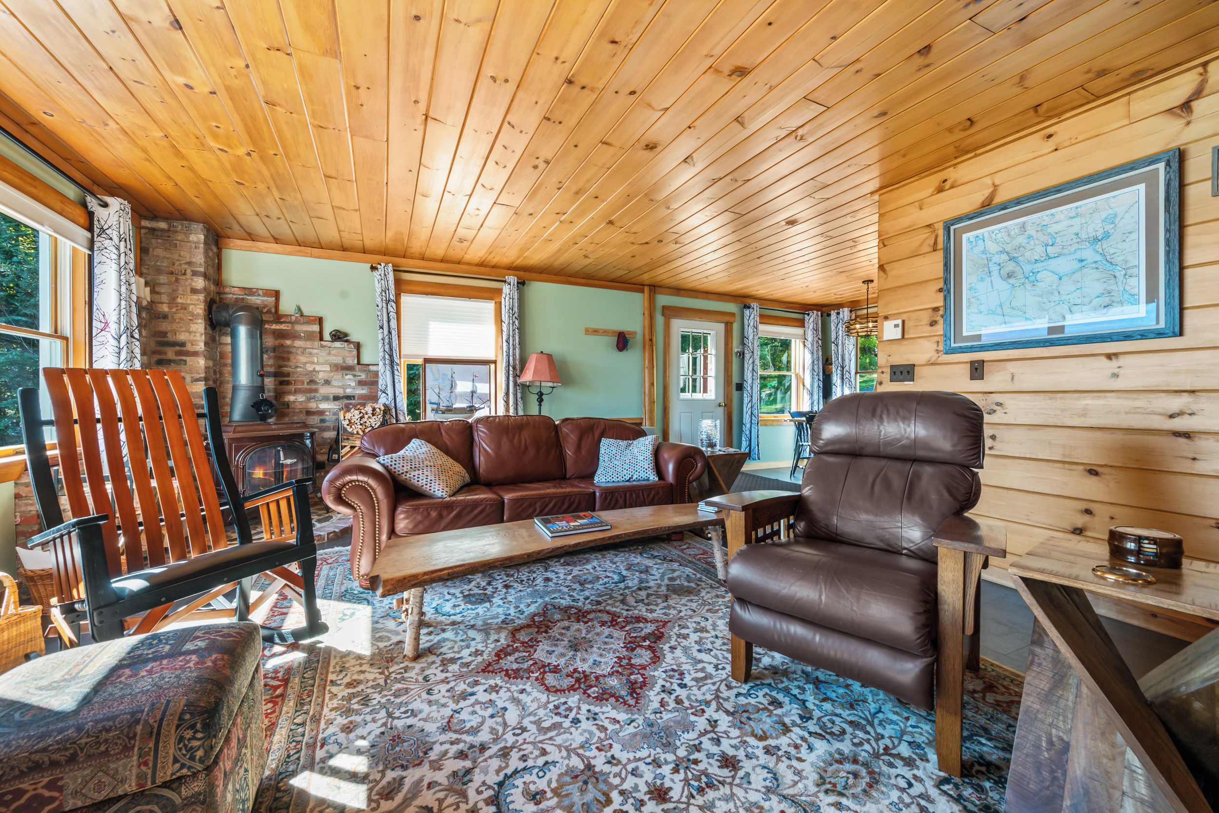 A cozy cabin interior with a wooden ceiling, a leather sofa, a wooden armchair, a patterned rug, and a fireplace.