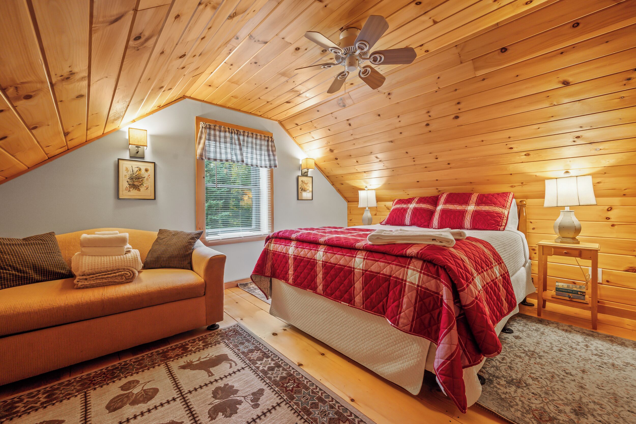 Une chambre mansardée chaleureuse avec un plafond en pente, des murs en bois, un lit recouvert d’une courtepointe rouge, un canapé avec une couverture et deux lampes fixées au mur.