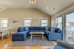 A living room with a sectional sofa, a coffee table, and a patterned rug. There are framed pictures on the wall, a ceiling fan, and large windows.
