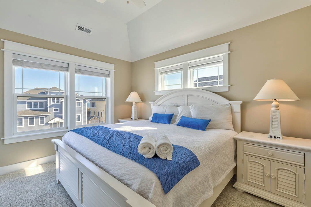 Une chambre à coucher avec un grand lit, deux tables de chevet, une commode et une vue sur les maisons par les fenêtres.
