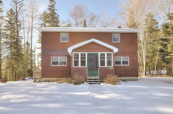 A two-story wooden cabin with a green door, surrounded by snow and trees.