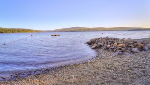 A pebbly beach with a wooden dock extending into calm waters, surrounded by hills and clear skies. - Rangeley Manor P | 4 Bedroom Cabin with Shared Waterfront Access (Rangeley)