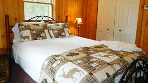 A wooden cabin bedroom with a bed, a nightstand, a lamp, and a door. - Rangeley Manor P | 4 Bedroom Cabin with Shared Waterfront Access (Rangeley)
