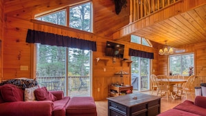 A cabin interior with a large window, a dining area, and a TV mounted on the wall. - Mahara 2 - Spacious home with access to Rangeley Lake and the snowmobile trails. (Rangeley)