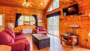 A cozy cabin interior with a wooden ceiling, a television mounted on the wall, a red sofa, a wooden coffee table, and a large window with curtains. - Mahara 2 - Spacious home with access to Rangeley Lake and the snowmobile trails. (Rangeley)