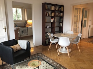 A dining area with a round table, white chairs, a bookshelf, and a kitchen in the background.