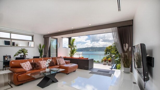 A modern living room with a leather sofa, a coffee table, and a large TV. There is a balcony with a view of the sea and mountains. - Villa 29/6 Ocean front pool villa (Tambon Patong)