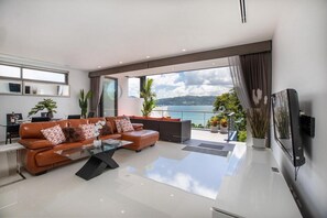 Una sala de estar moderna con un sofá de cuero, una mesa de centro y un televisor grande. Hay un balcón con vista al mar y a las montañas.