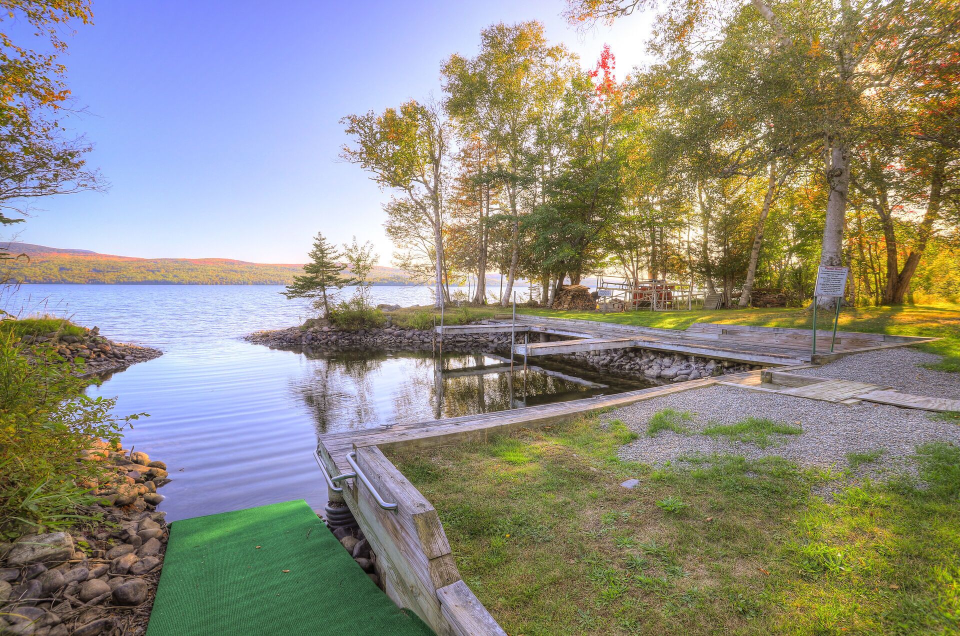 A serene lakeside dock with a wooden walkway leading to a grassy area, surrounded by trees and a clear sky.