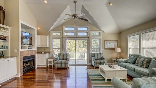 A spacious living room with hardwood flooring, a fireplace, and a large window overlooking a deck. - Jones Beach House | 1200 ft from the beach | Private Pool, Hot Tub | Corolla (Corolla)