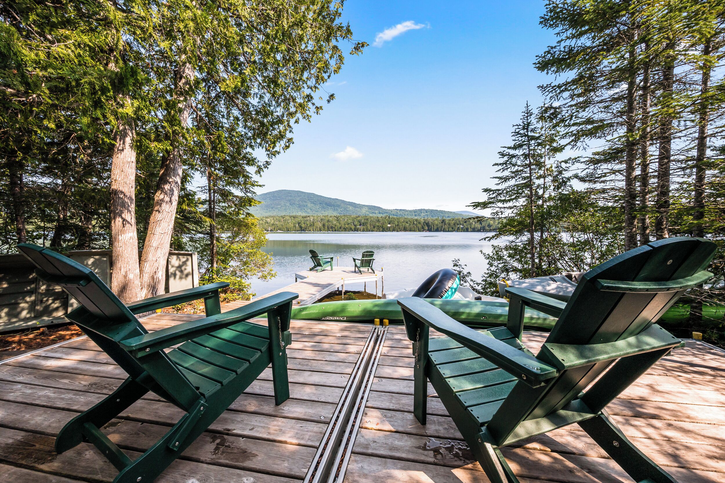 A deck with green Adirondack chairs overlooking a lake, surrounded by trees.