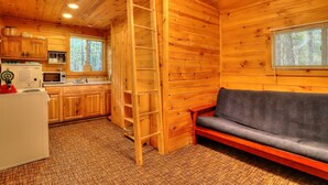 L’intérieur d’une cabane avec des murs en bois, un coin cuisine, un canapé et une échelle.
