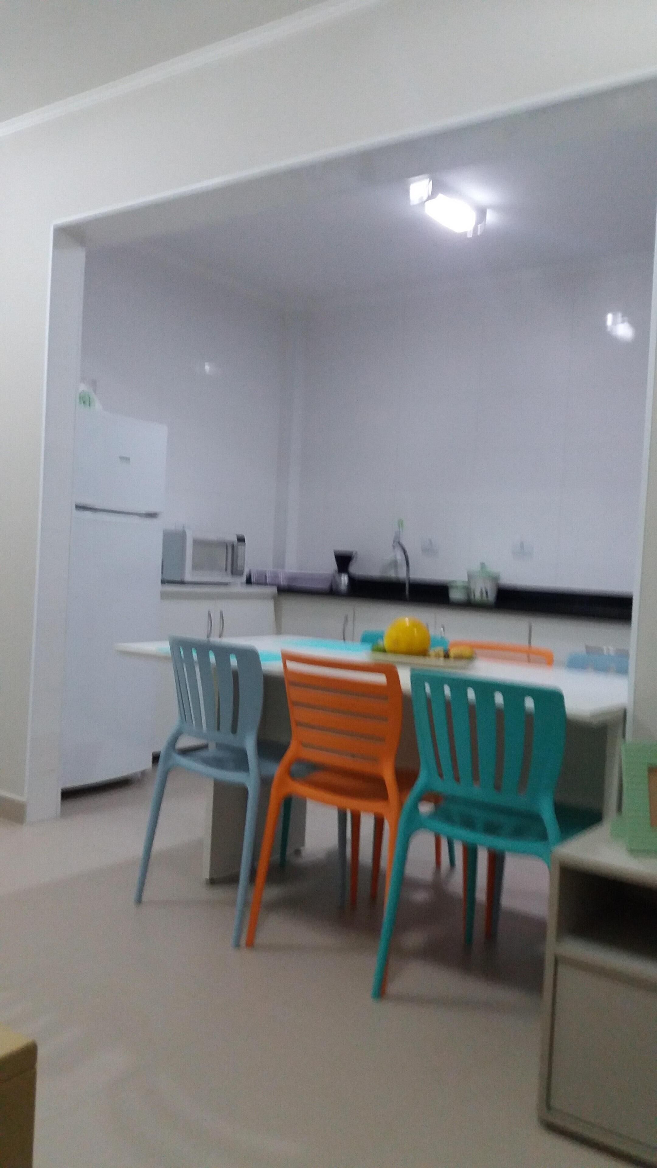 A modern kitchen with white cabinets, a white refrigerator, a microwave, and a sink. There are colorful chairs around a table with a yellow fruit bowl.