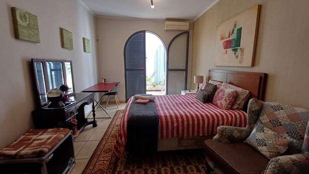 Ein Schlafzimmer mit einem Bett, einem Schreibtisch, einem Sessel, einer Couch und einem kleinen Tisch. Ein Fenster und eine Klimaanlage sind an der Wand angebracht.