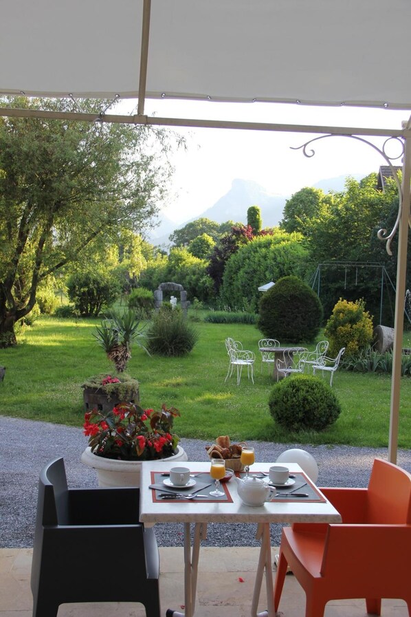 An outdoor dining area with a table set for breakfast, surrounded by chairs, and overlooking a lush garden with a mountain in the background.