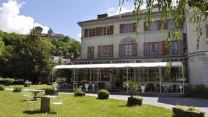 Un bâtiment à deux étages doté d’un espace extérieur aménagé avec des sièges, le tout entouré de jardins et d’arbres bien entretenus.