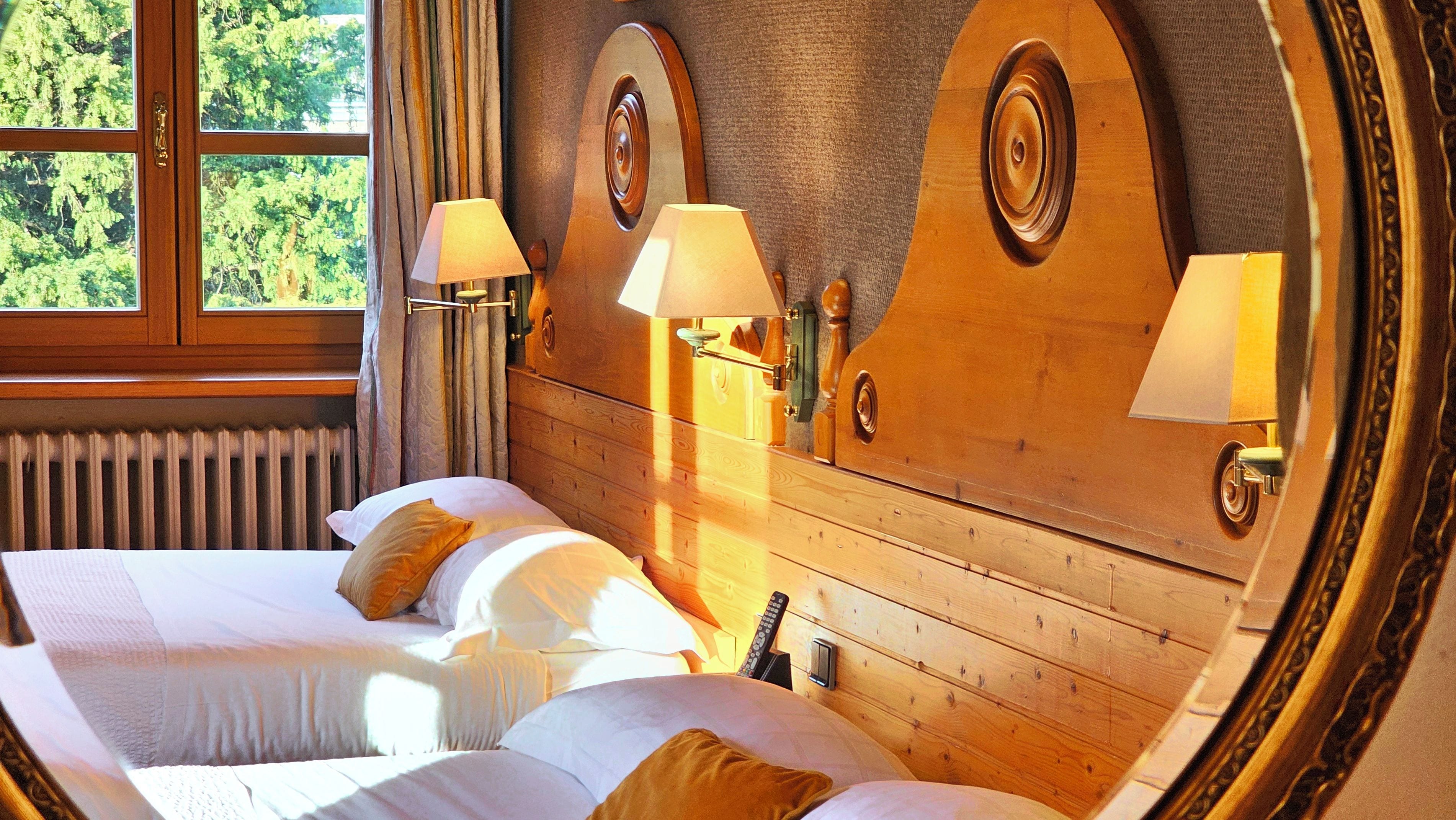 A hotel room with a wooden headboard, two bedside lamps, a window with curtains, and a bed with pillows.
