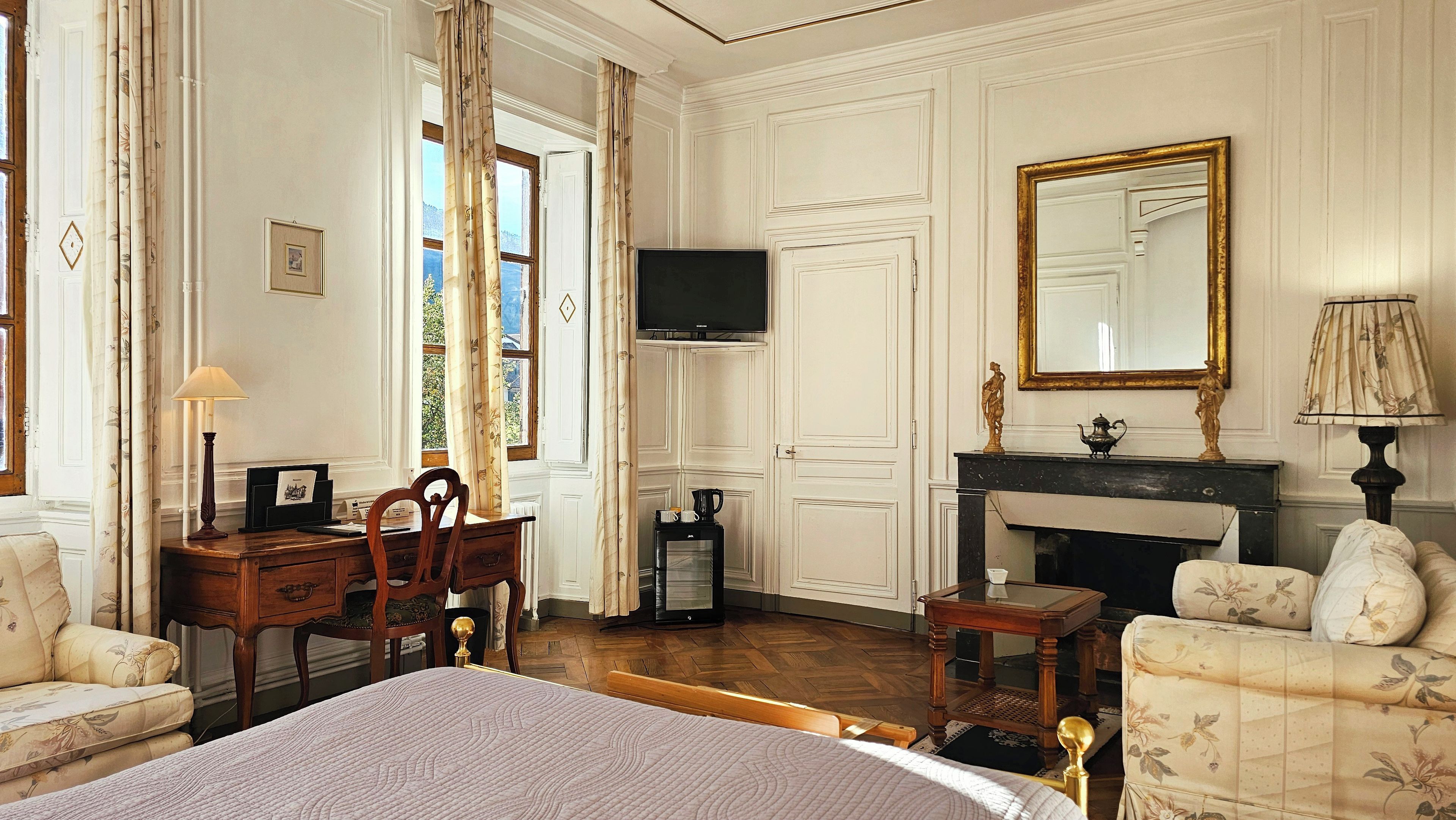 A classic hotel room with a bed, a desk, a television, a fireplace, and a large mirror.