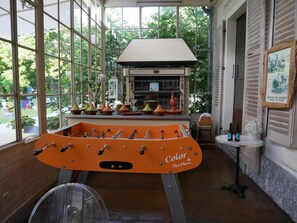 A room with a foosball table, a window with a view of greenery, and a table with bottles and a vase. - Hotel du Parc - Manoir du Baron Blanc (Faverges-Seythenex)