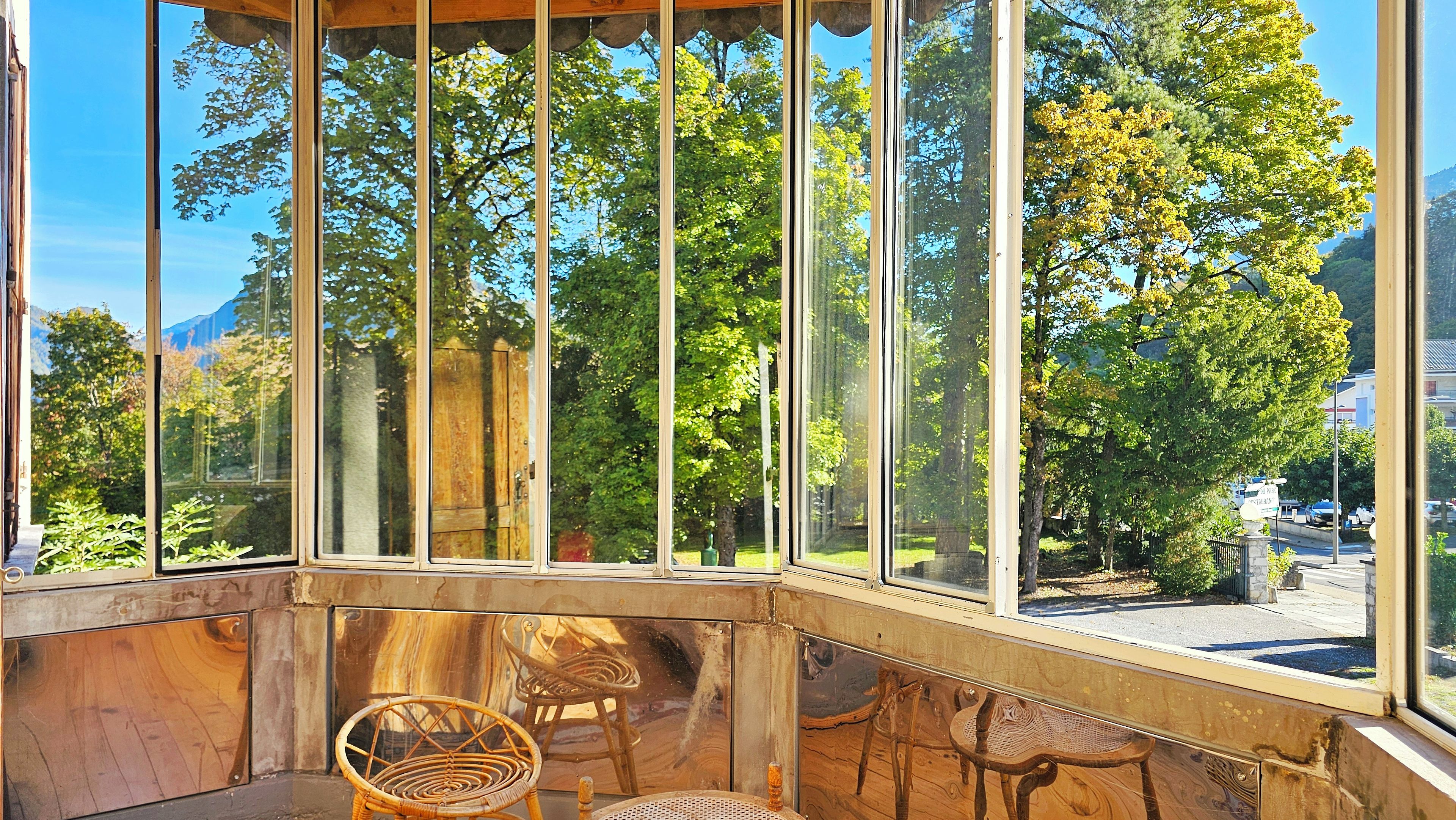 Ein Wintergarten mit großen Fenstern, Korbmöbeln und Blick auf Bäume und Berge.