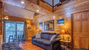 A cozy cabin interior with a wooden ceiling, a sofa, a dining table with chairs, and a small side table with a lamp and decorative items. - 27 Depot - Adorable 2 bedroom, 2 bath cottage close to the village (Rangeley)