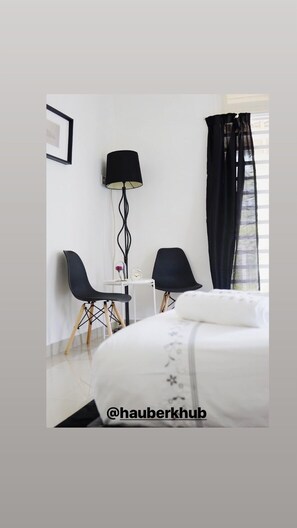 Ein Hotelzimmer mit einem Bett, zwei Stühlen, einem kleinen Tisch mit einer Uhr, einer Lampe und einem Fenster mit Vorhängen.