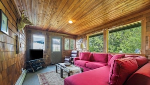 Ein Holzblockhaus-Innenraum mit einem roten Sofa, einem Fernseher, einem Ledersessel und einem großen Fenster mit Blick auf Bäume.