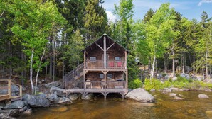 A cabin with a wooden deck and a balcony, surrounded by a forest and a river. - Rock Ledge - Relax & Renew in turn-of-century log cabin with side cabin (Rangeley)