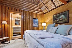 A wooden cabin bedroom with a bed, a nightstand, a lamp, a framed picture, and a door leading to a bathroom.