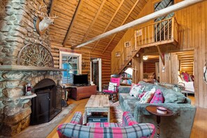 A log cabin interior with a stone fireplace, wooden ceiling, a television, a fireplace mantel, a sofa, and a bookshelf.