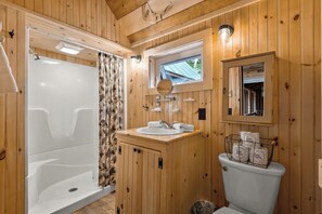 A wooden cabin bathroom with a shower, toilet, sink, and a window above the mirror.