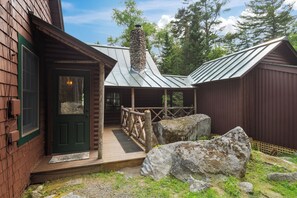 A rustic cabin with a stone chimney, wooden deck, and a metal roof.
