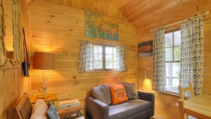A cozy cabin interior with wooden walls, a sofa with a cushion, a table, and two windows with patterned curtains. - Greenvale Cove 8 - Greenvale Cove cottage community on Rangeley Lake (Rangeley)
