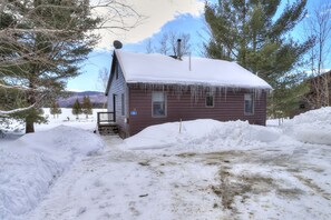 A wooden cabin with a snow-covered roof and a satellite dish, surrounded by snowbanks and trees. - Saddleback Lake Lodge 33 | Close to Saddleback (Rangeley)