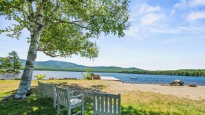 A lakeside view with two wooden benches under a tree, a dock, and a calm lake with mountains in the background. - Saddleback Lake Lodge 33 | Close to Saddleback (Rangeley)