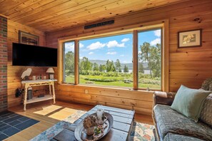 Ein gemütliches Blockhaus-Interieur mit einem großen Fenster, das einen Blick auf einen See und Grünflächen bietet.